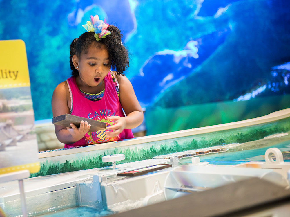 Child holding a toy boat with her mouth wide open as she gazes into a water table.